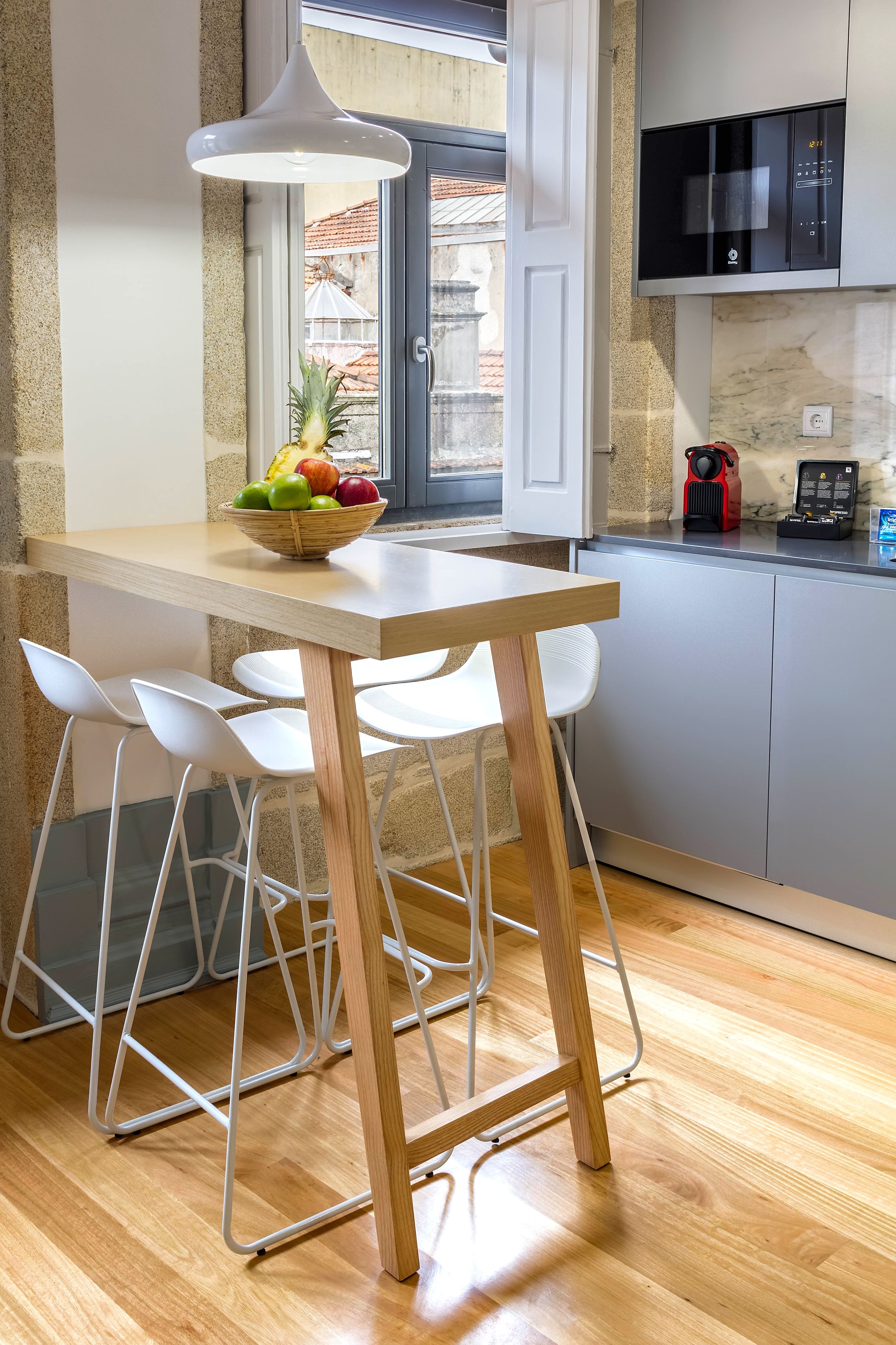 Une cuisine compacte avec une table en bois, des chaises blanches, un panier de fruits, un four à micro-ondes et une machine à café.