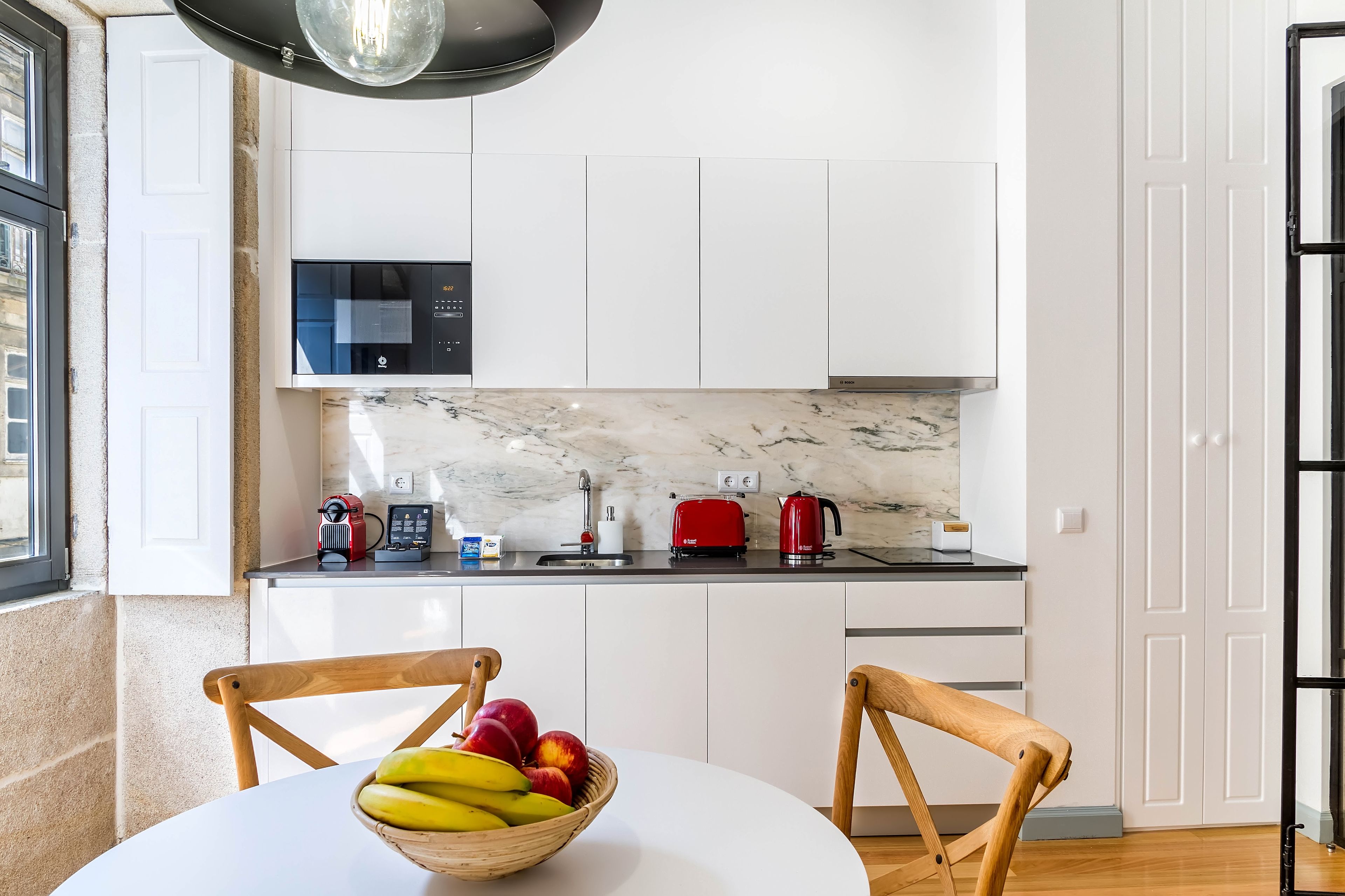 A modern kitchen with white cabinets, a marble countertop, a microwave, a red toaster, and a kettle. There is a wooden chair and a table with a fruit bowl.