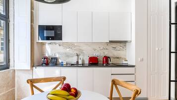 A modern kitchen with white cabinets, a marble countertop, a microwave, a red toaster, and a kettle. There is a wooden chair and a table with a fruit bowl.