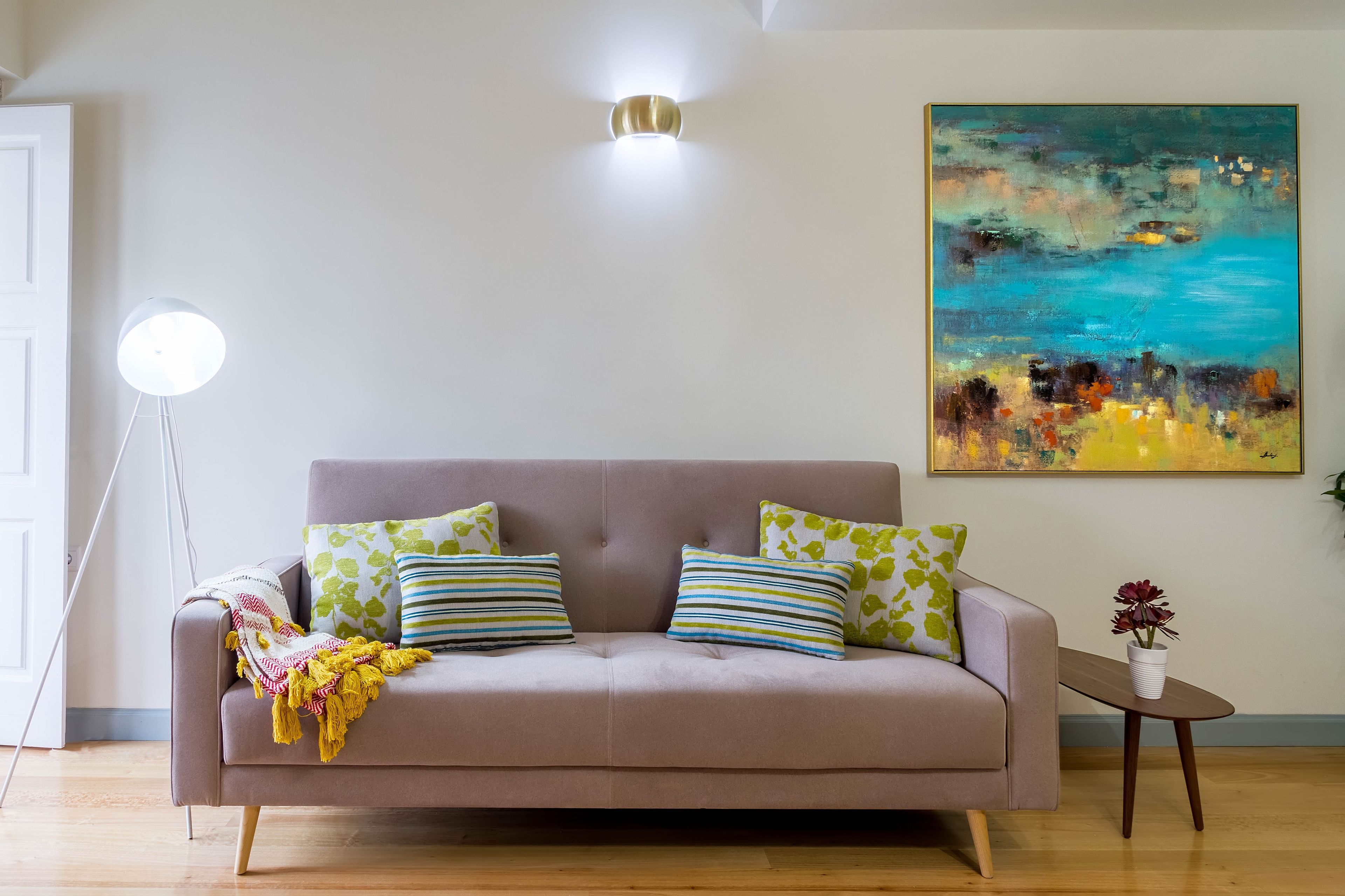 A living room with a sofa, pillows, a floor lamp, a painting, and a small table with a plant.