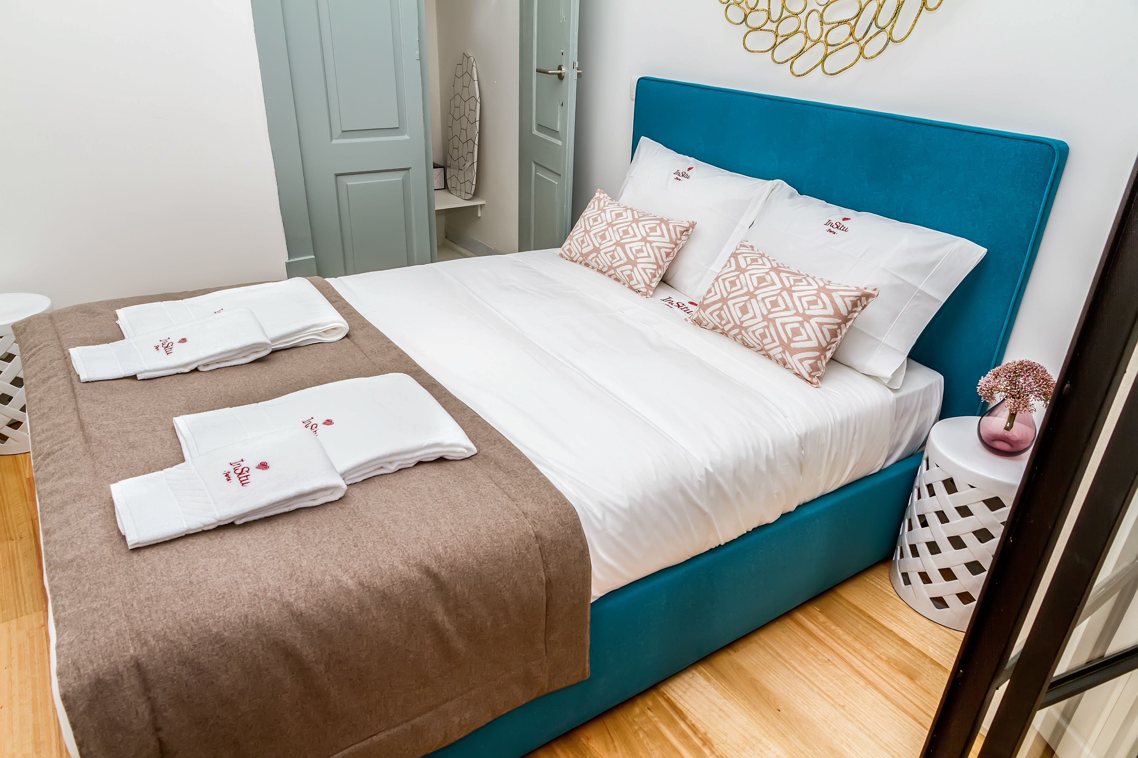 Una cama bien hecha con almohadas blancas y estampadas, un plaid marrón y toallas dobladas con el logotipo de un corazón.