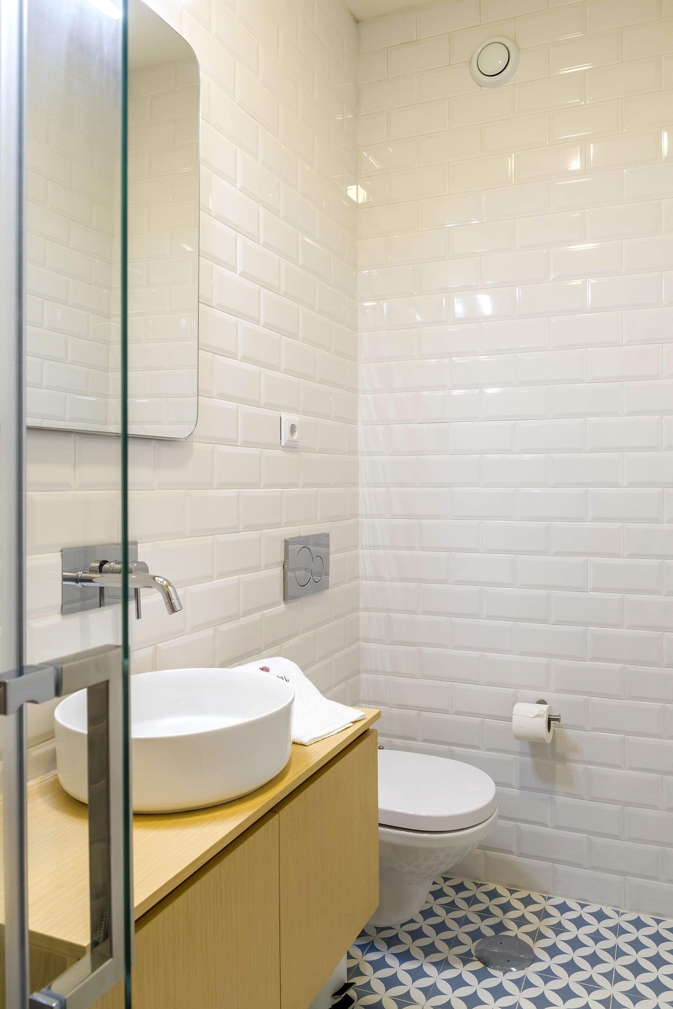 Un baño moderno con azulejos blancos tipo metro, un lavamanos redondo, un inodoro y un piso con diseño.