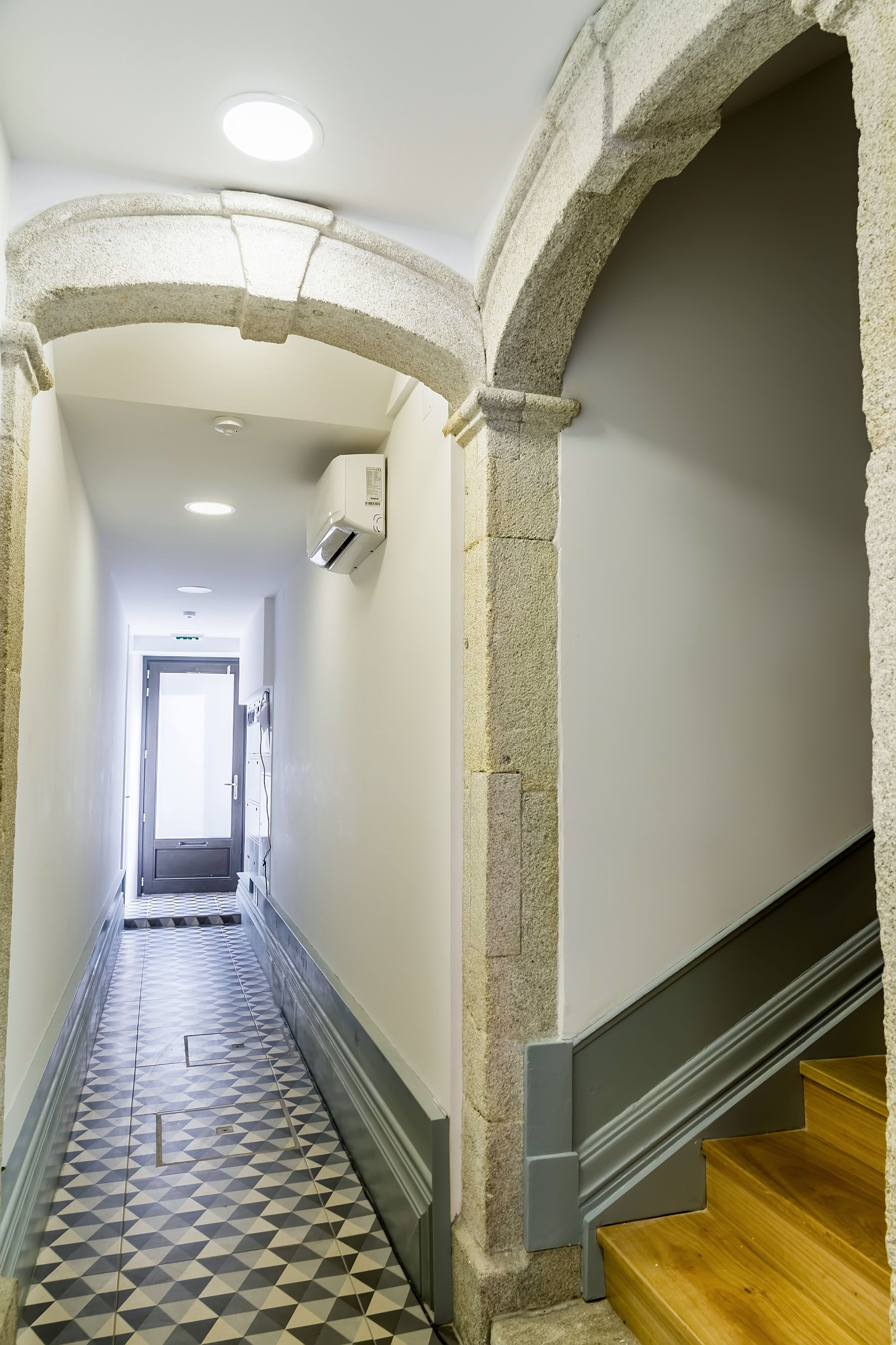A narrow hallway with a geometric patterned floor, stone arches, and a wooden staircase.