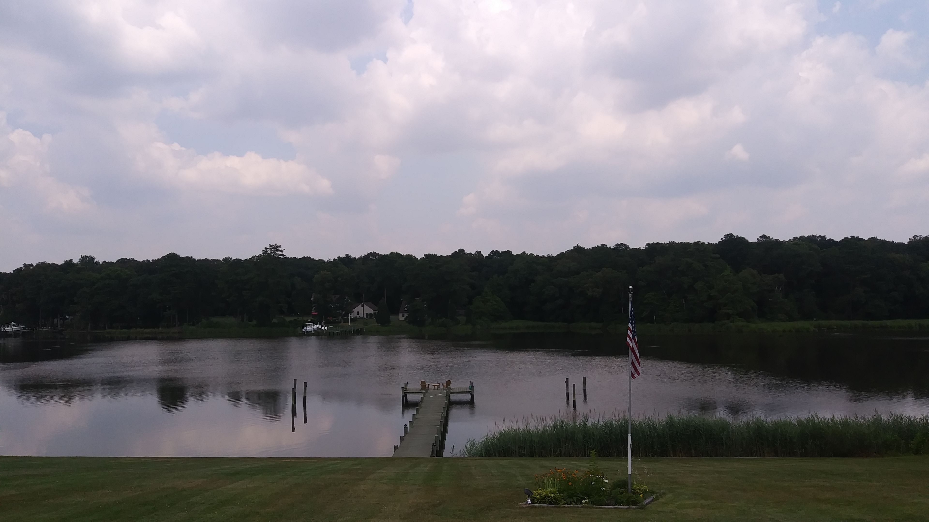 A calm lake with a dock extending into the water, surrounded by lush greenery and a forested hill in the background.