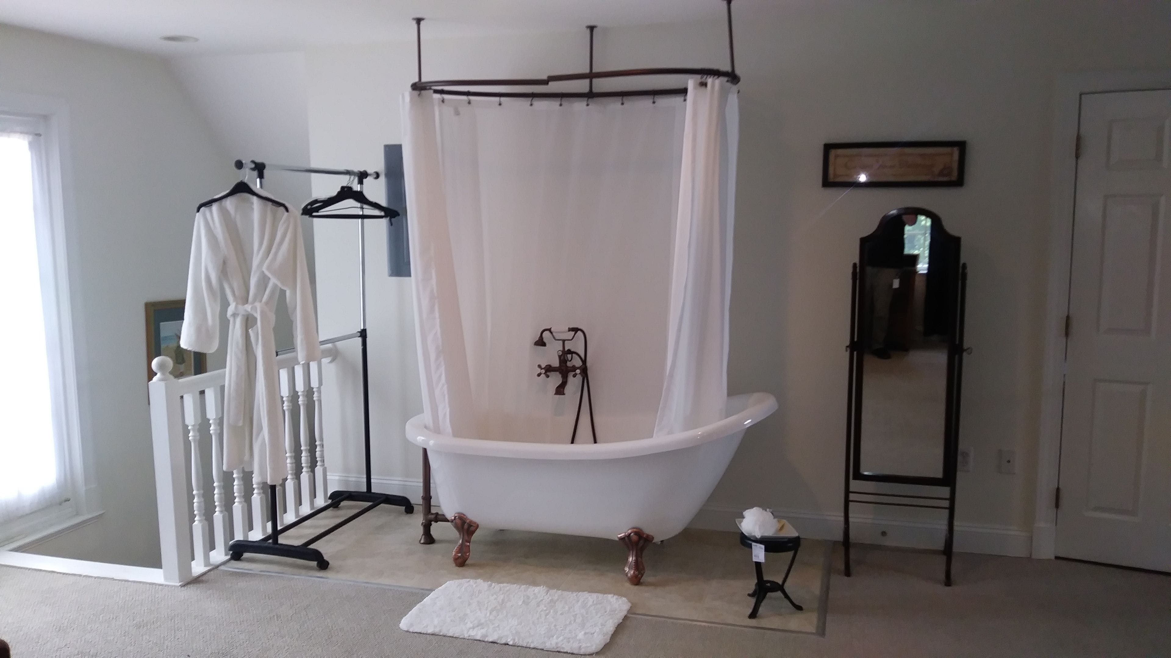 Un baño con una bañera independiente, una cortina de ducha blanca, un perchero con toallas, una mesita con una caja de pañuelos y un espejo en la pared.