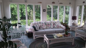A sunlit room with a checkered sofa, wicker chairs, a glass table, and large windows offering a view of greenery. - Marvel's on the Creek (East New Market)
