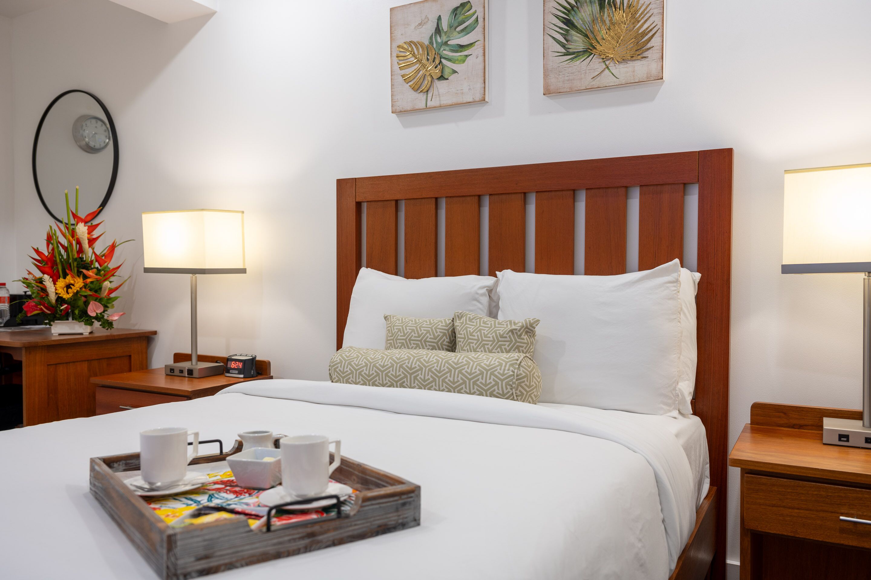 A neatly made bed with a wooden headboard, two bedside lamps, a tray with cups and a book, and a clock on the wall.