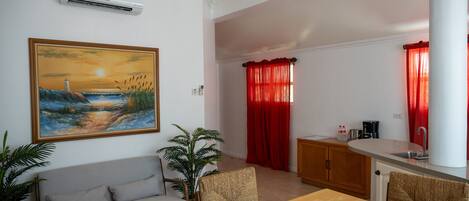 Una sala de estar con sofá, una mesa de comedor y sillas, una cocina con pileta y armarios, y un cuadro en la pared.