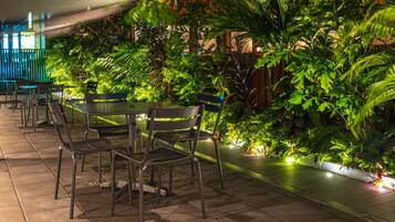 An outdoor patio with tables and chairs, surrounded by lush greenery and a wooden fence.