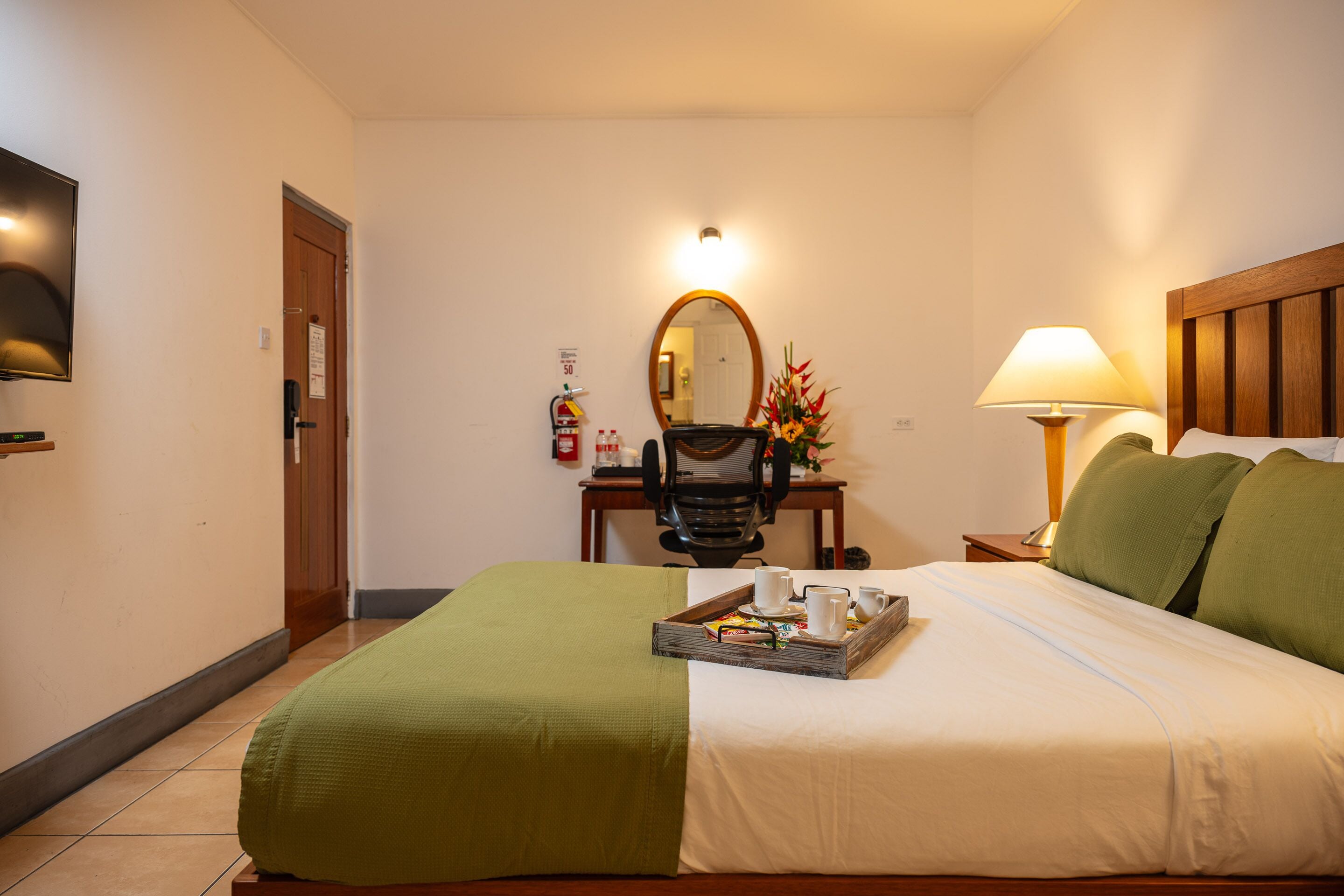 A hotel room with a bed, a desk with a chair, a mirror, a lamp, and a tray with breakfast items.
