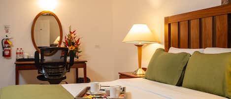 A hotel room with a bed, a desk with a chair, a lamp, a mirror, and a tray with cups and a flower arrangement.