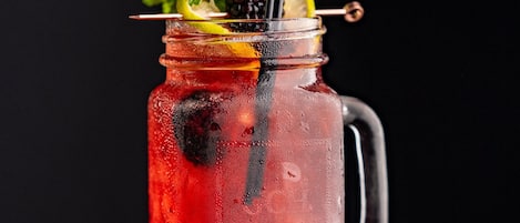 Un frasco de vidrio con una bebida roja, adornado con zarzamoras, menta y una rodaja de limón, sobre una superficie de madera.