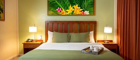 Una habitación de hotel con una cama grande, dos mesitas de noche con lámparas, una manta verde y una bandeja con una taza y un libro.