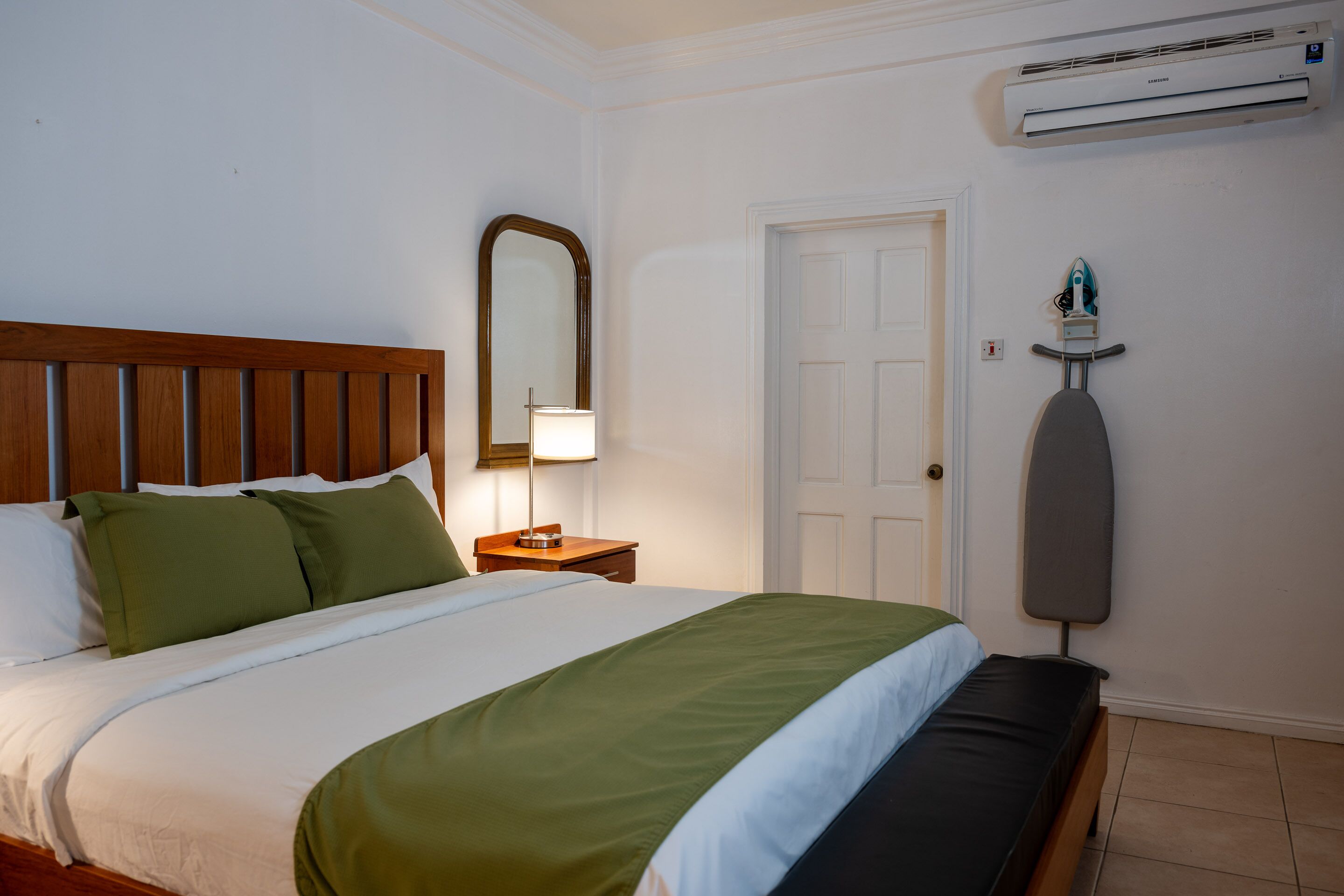 A neatly made bed with green and white bedding, a wooden headboard, a bedside lamp, and an ironing board against the wall.