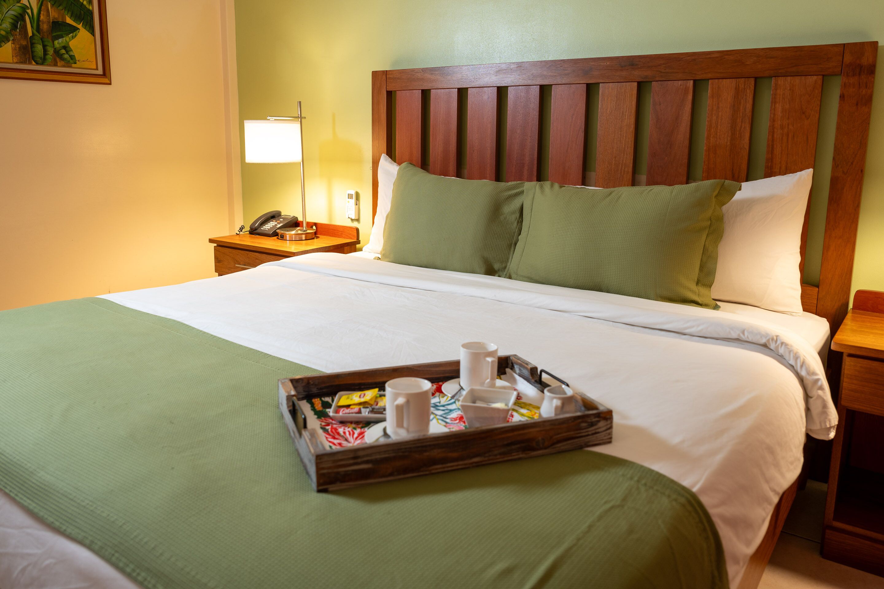 Una cama bien tendida con cabecera de madera, una mesita de noche con lámpara y una bandeja con alimentos para el desayuno.