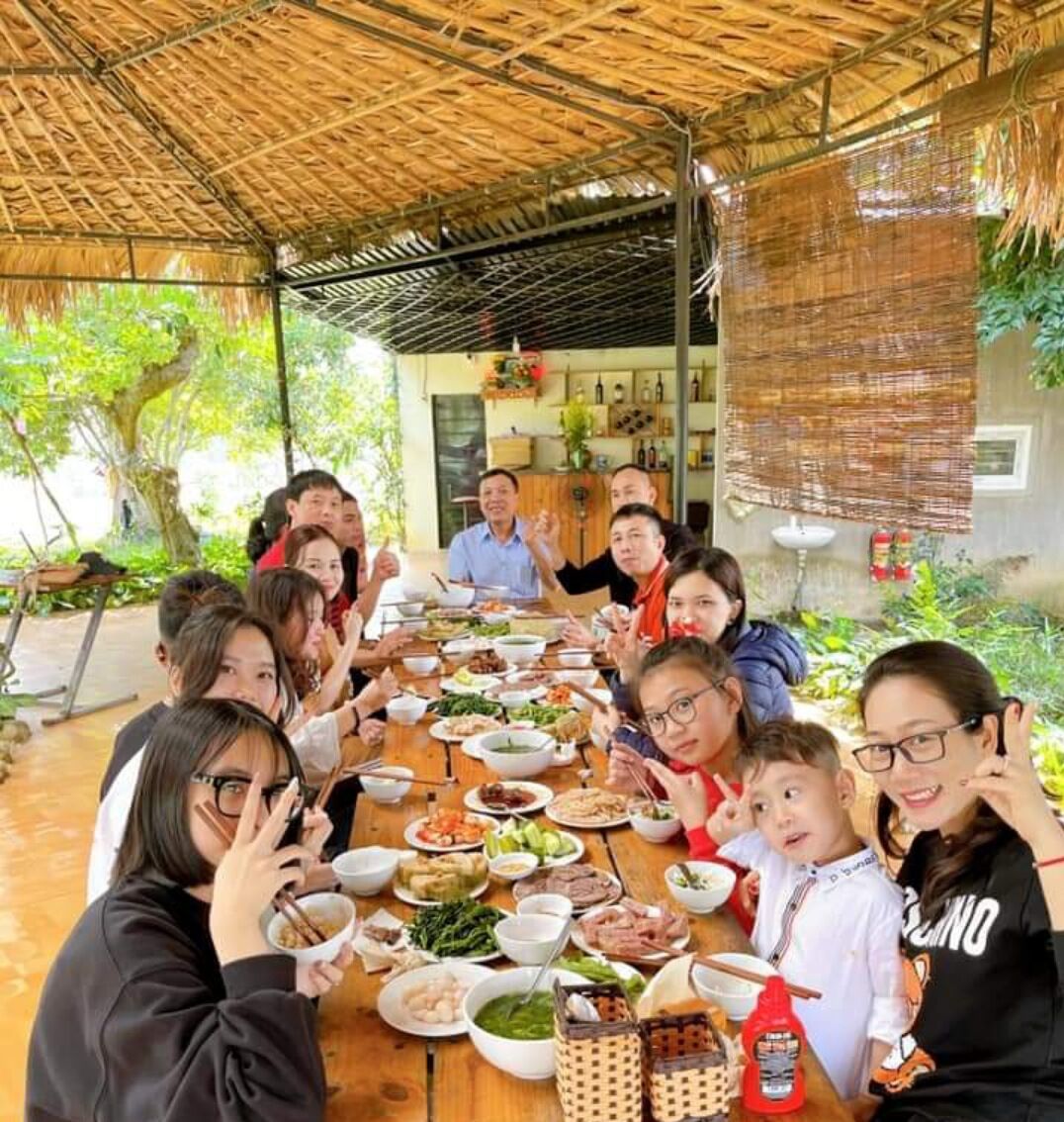 A group of people seated around a wooden table with various dishes, under a thatched roof structure.