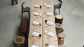 Une longue table en bois, dressée pour le repas, avec des chaises et des tabourets disposés autour.