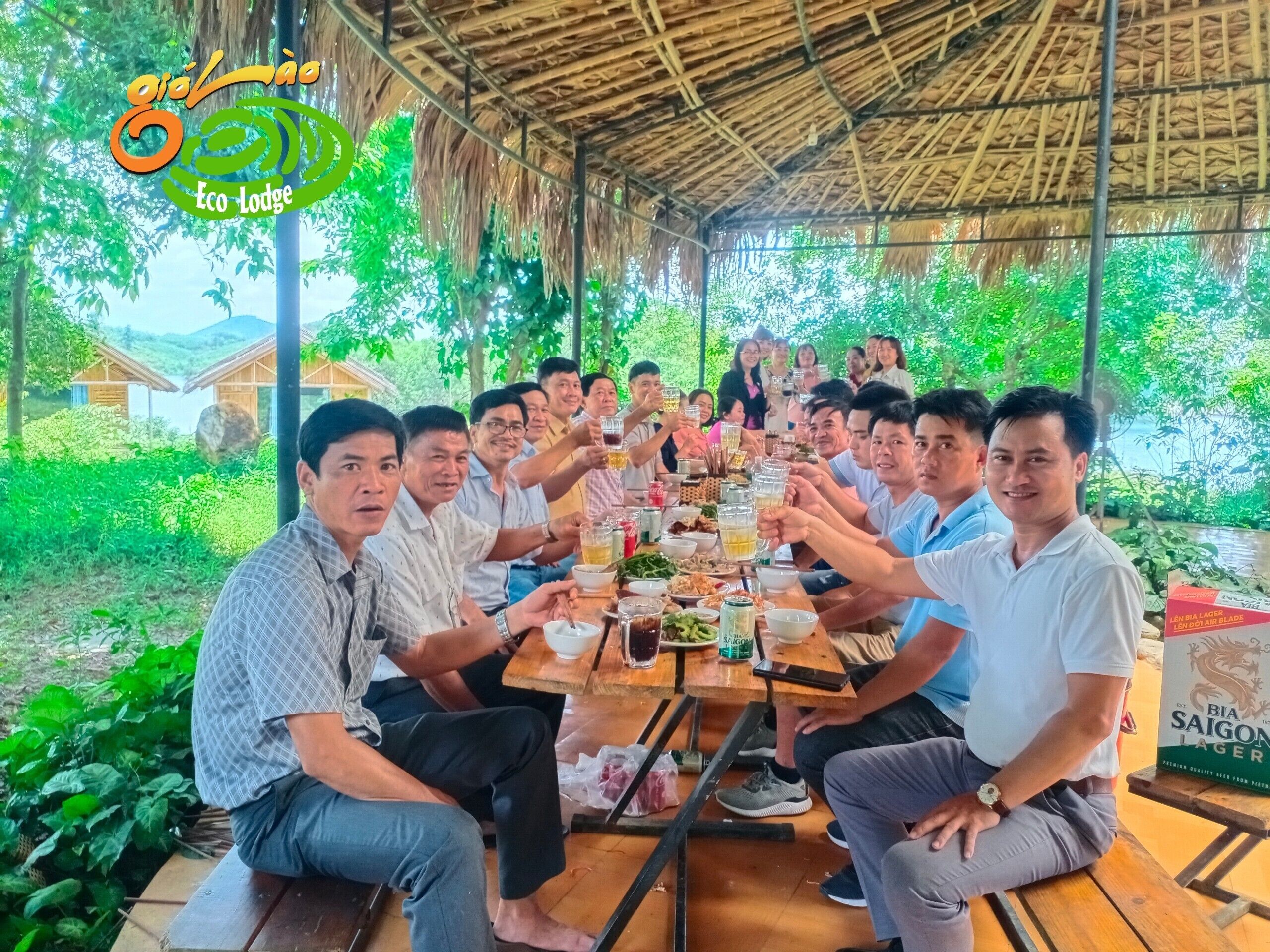 Un groupe de personnes est assis autour d’une table en bois à l’extérieur, dans un endroit verdoyant et abrité par une structure à toit de chaume.