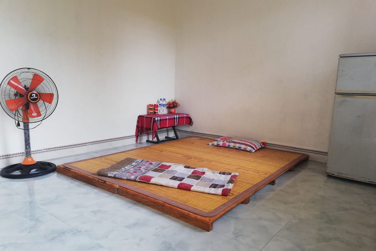 Ein Zimmer mit einem hölzernen Bett, einem rot-weiß karierten Tisch, einem Ventilator und einem Kühlschrank.