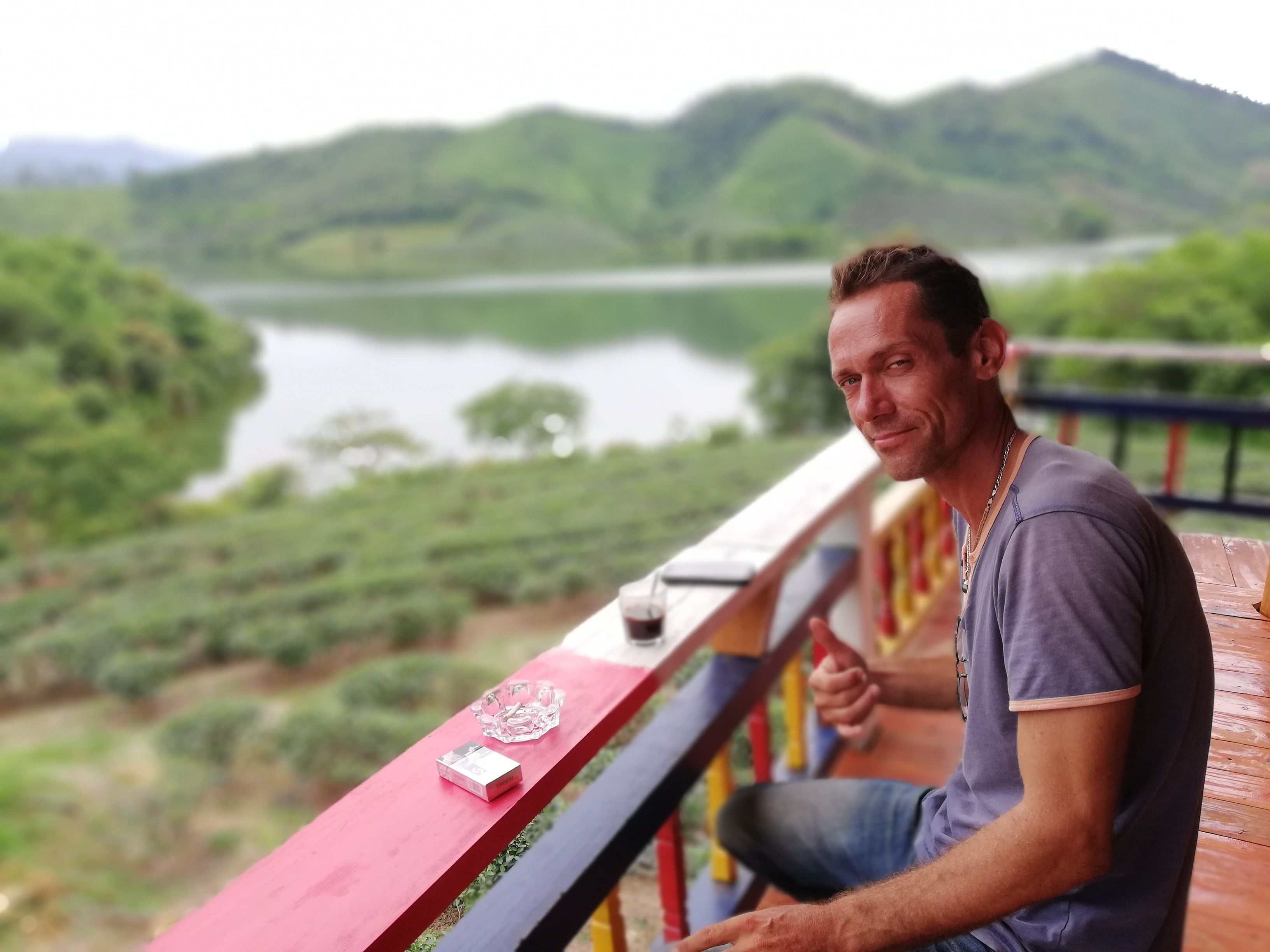 A man sitting on a wooden deck with a lake and hills in the background.