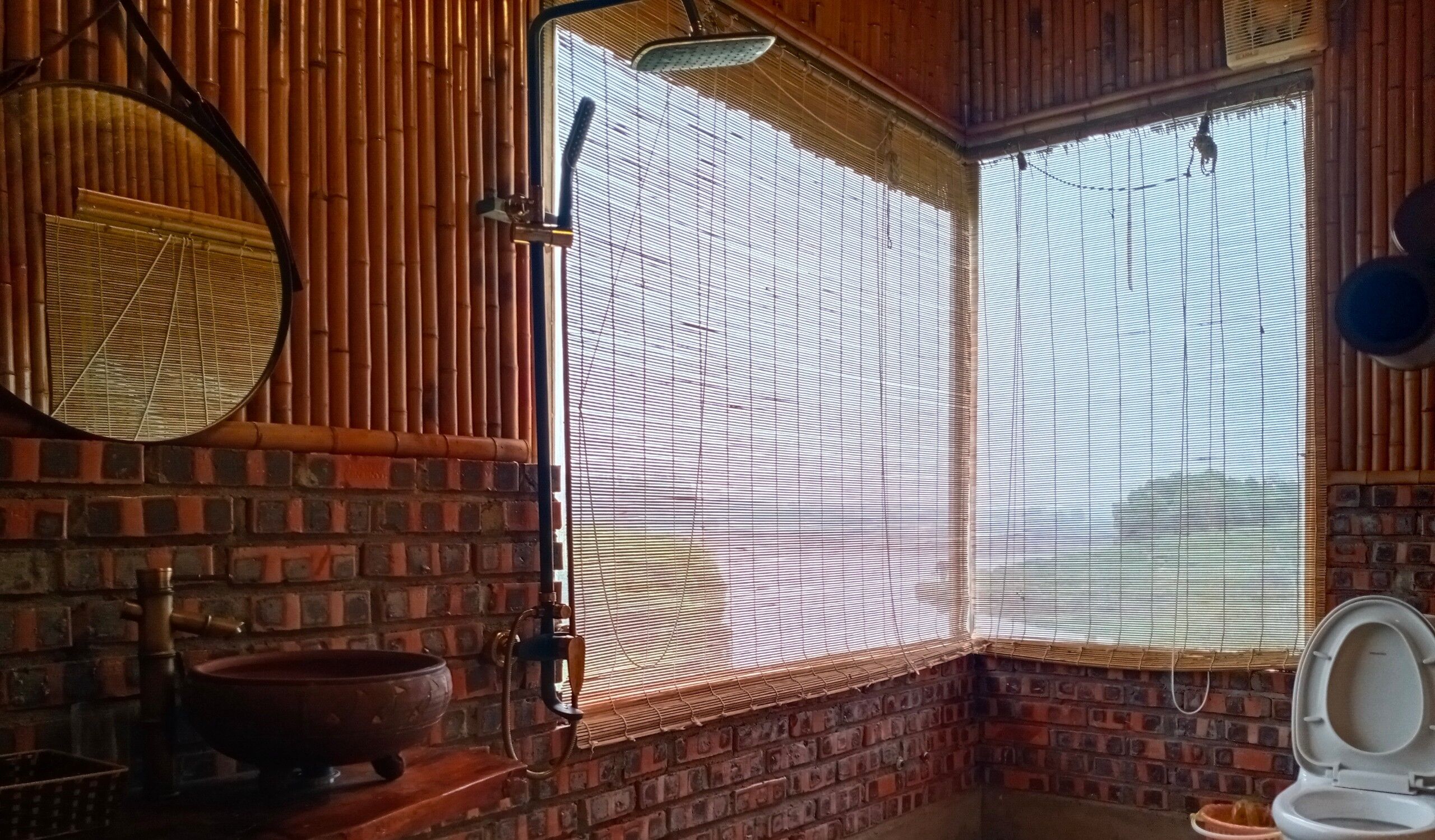 A bathroom with a brick wall, a round mirror, a traditional basin, and a window with bamboo blinds.