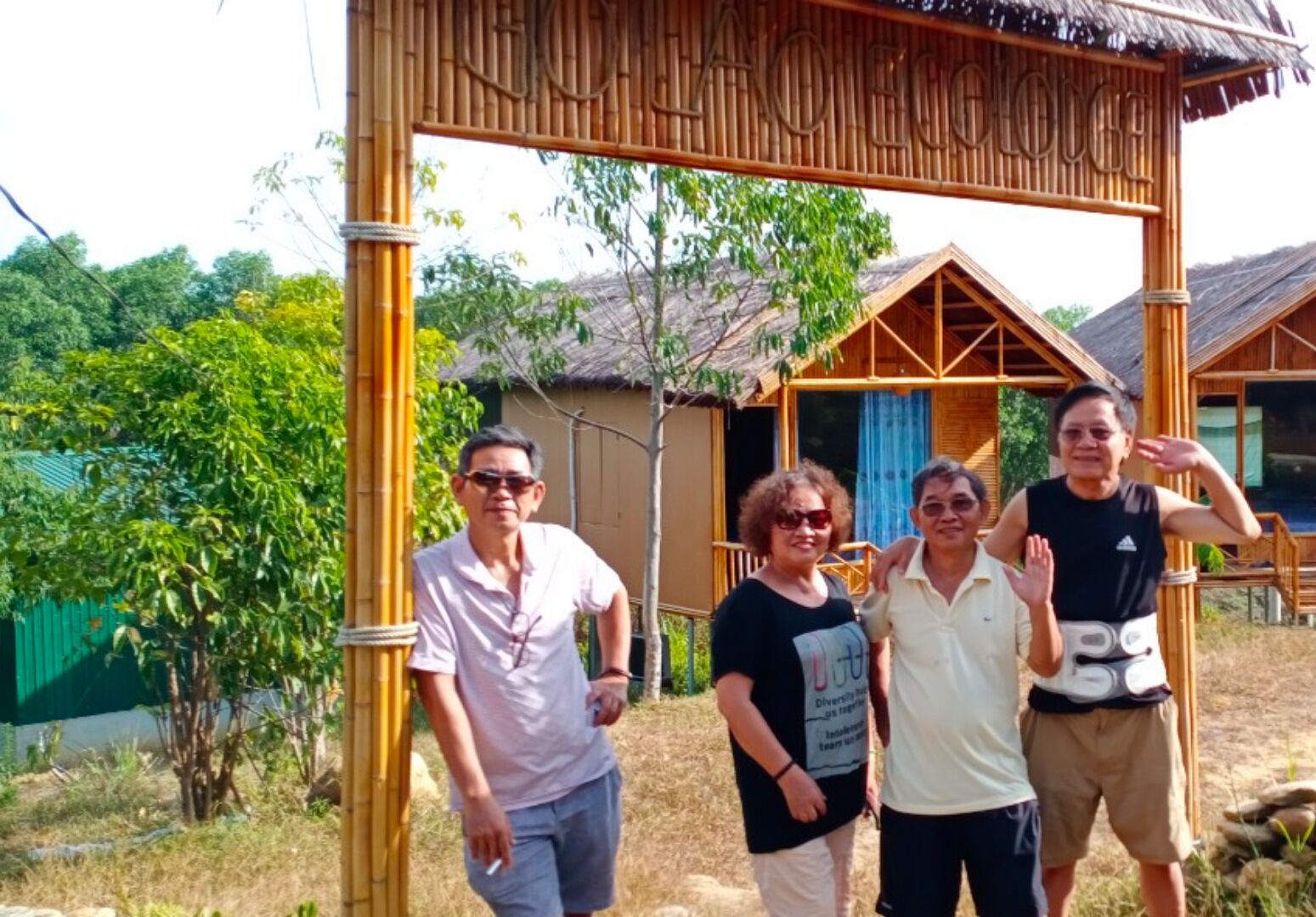 A bamboo structure with a thatched roof, surrounded by greenery and a building with a similar architectural style.