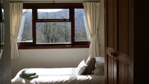 A bedroom with a bed, a window with curtains, and a view of trees and mountains. - Highgarry Cottage (Invergarry)