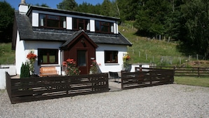 A two-story house with a white exterior and dark roof, a front porch with a bench, and a gravel driveway. - Highgarry Cottage (Invergarry)