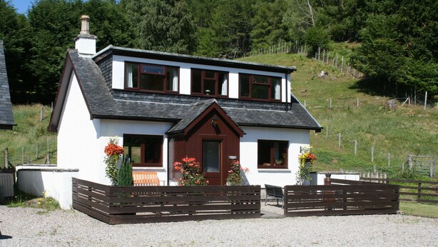 A two-story house with a white exterior and dark brown trim, featuring a front porch with wooden benches and flower boxes. - Highgarry Cottage (Invergarry)