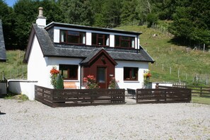 A two-story house with a white exterior and dark brown trim, featuring a front porch with wooden benches and flower boxes. - Highgarry Cottage (Invergarry)