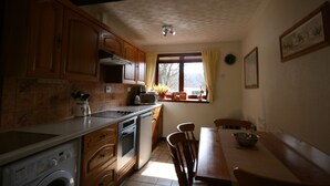A kitchen with wooden cabinets, a washing machine, a dining table with chairs, and a window with curtains. - Highgarry Cottage (Invergarry)