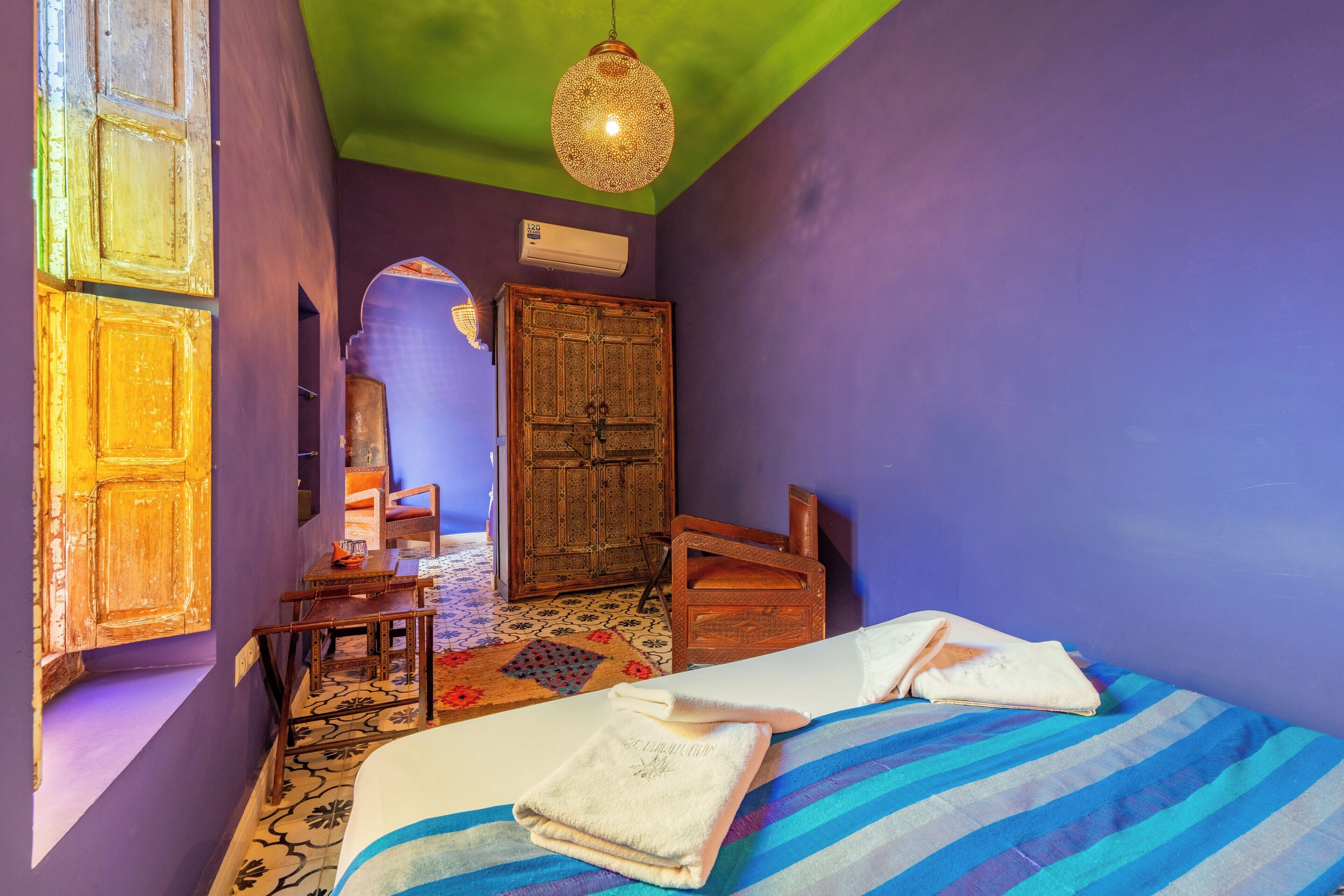 A room with a bed, a wooden wardrobe, a chair, a small table, and a hanging light fixture.