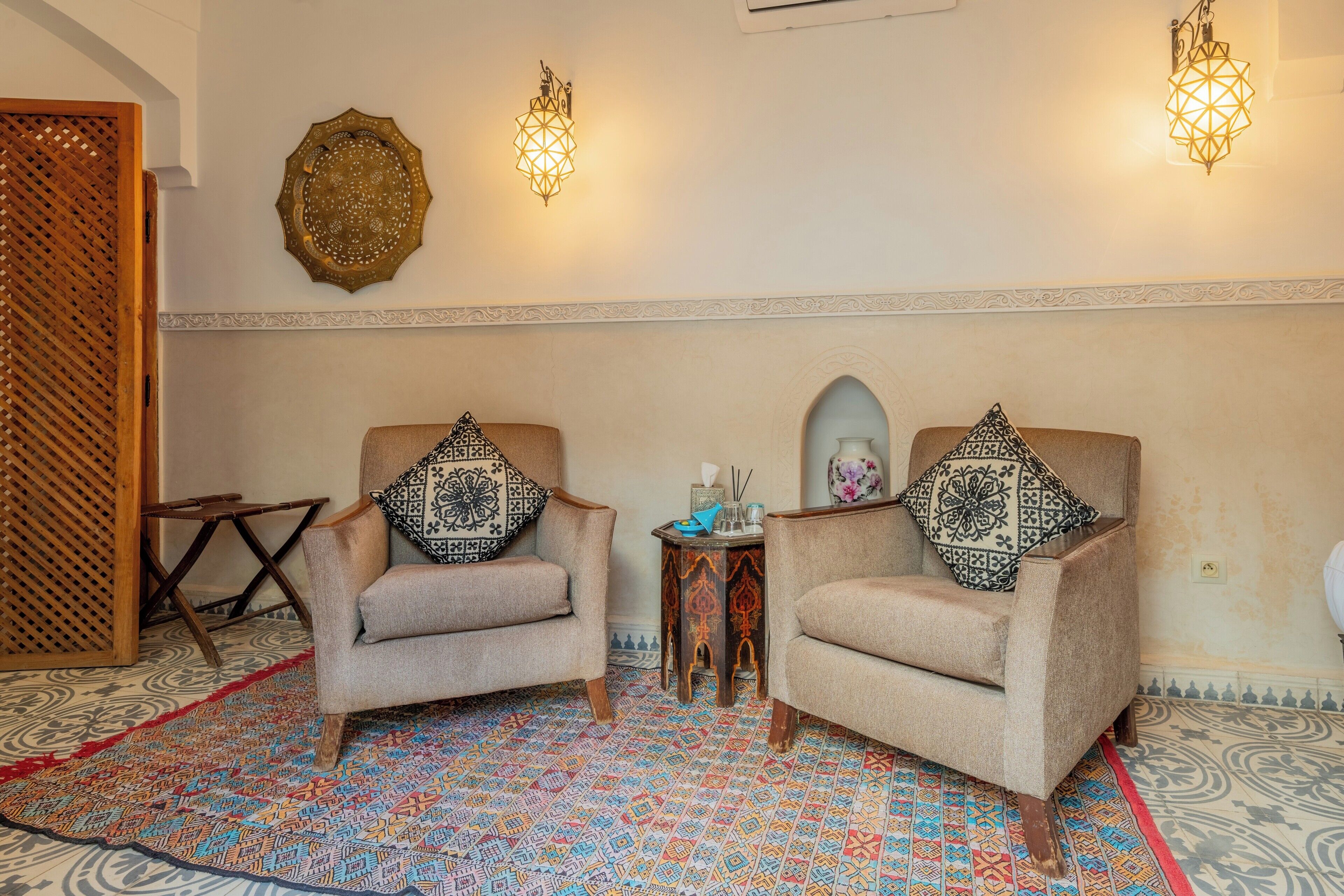 A room with two beige armchairs, a patterned rug, and decorative wall sconces.