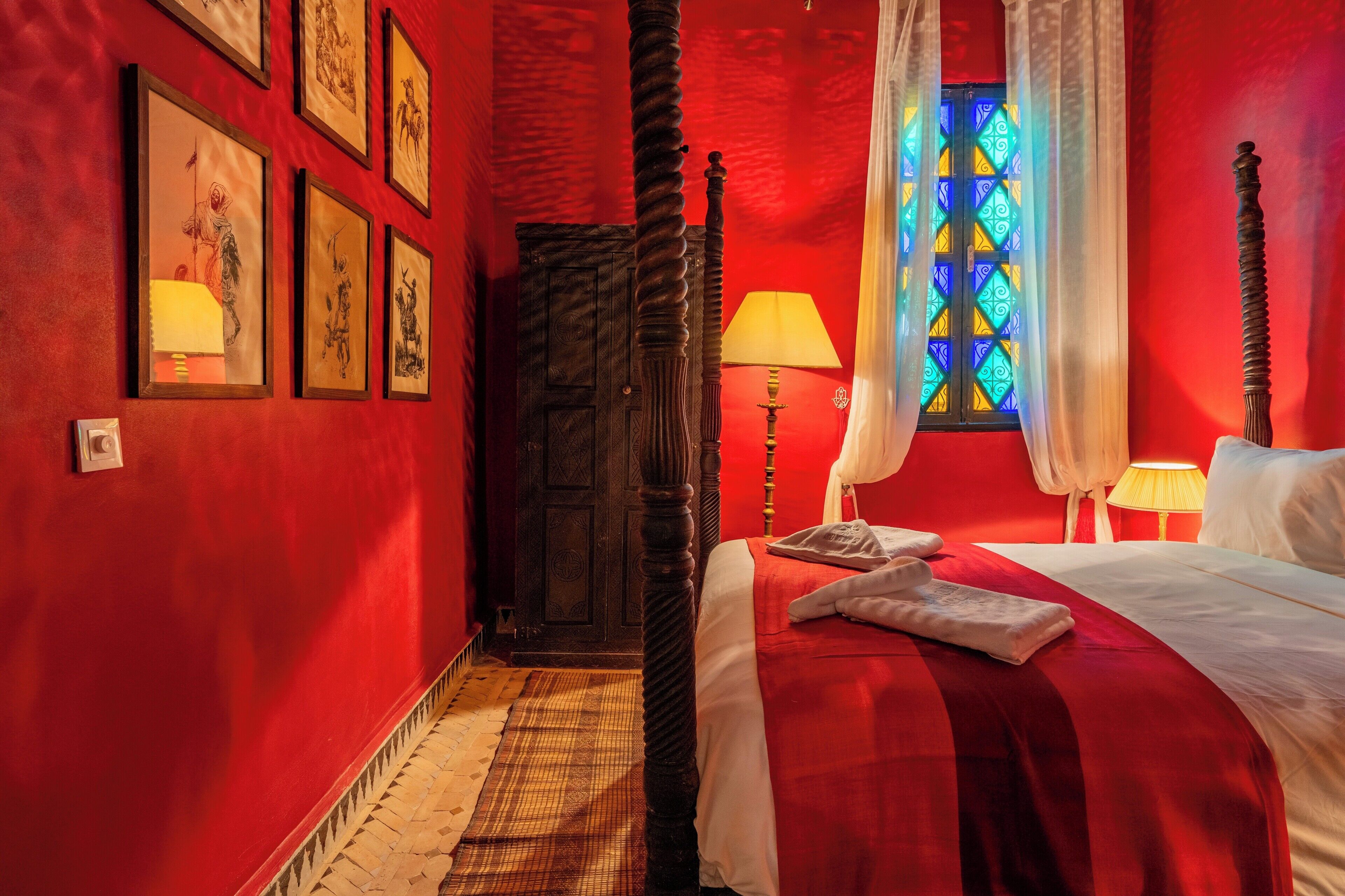 Una habitación con una cama, una mesita de noche, una lámpara, un ventanal con vidrieras y una puerta.