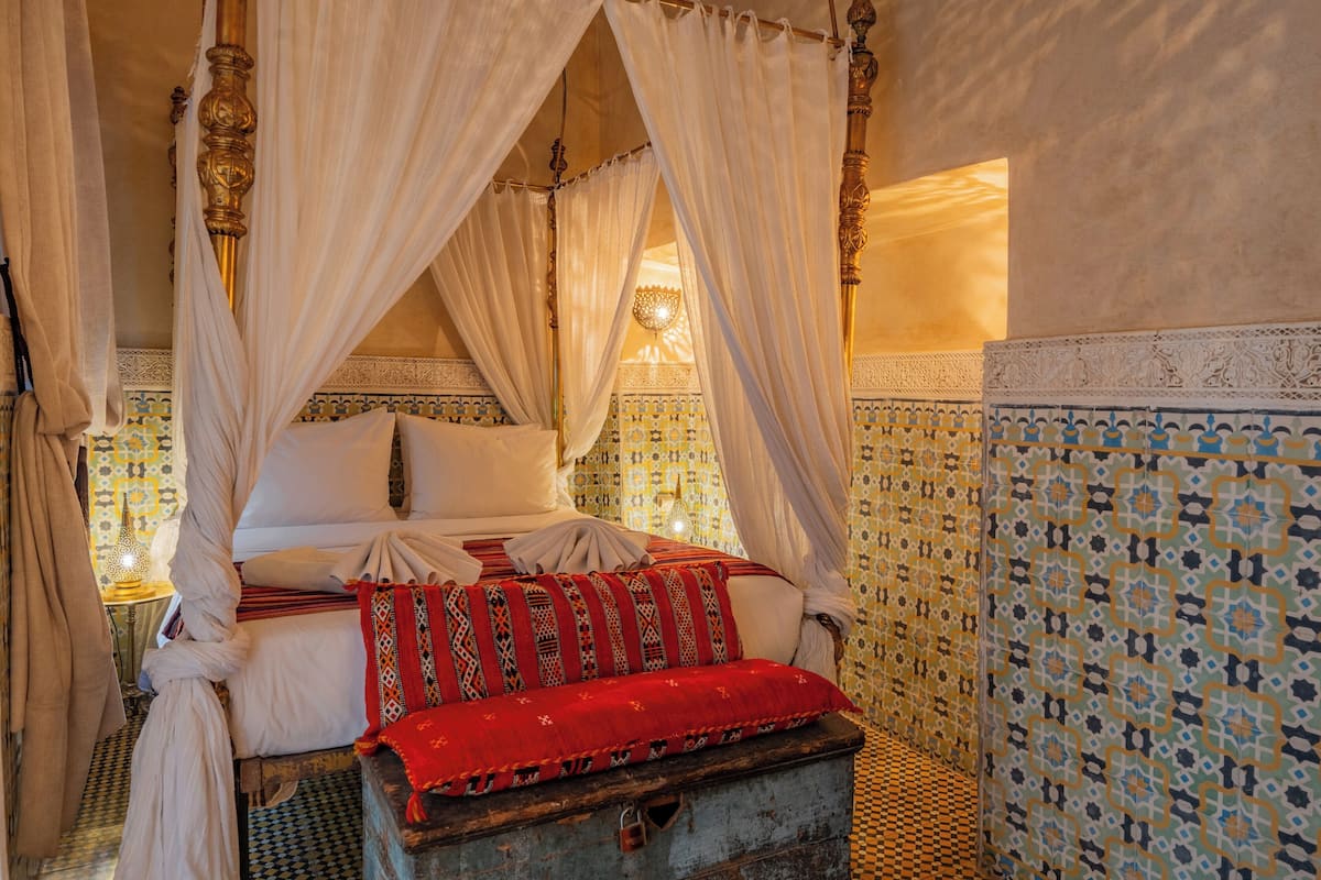 A four-poster canopy bed with white linens and a red patterned throw, set against a backdrop of blue and yellow patterned tiles.