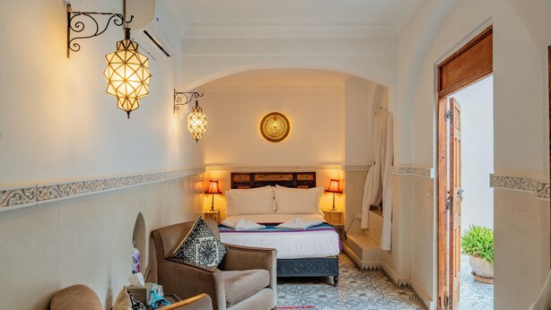 A narrow hallway leading to a bedroom with a bed, two bedside lamps, a decorative wall hanging, a potted plant, and a patterned rug. - Riad Yamina52 (Marrakech)