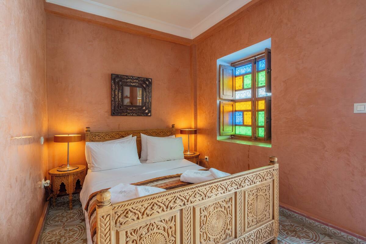 A room with a bed, two bedside lamps, a decorative mirror, and a window with colorful panes.
