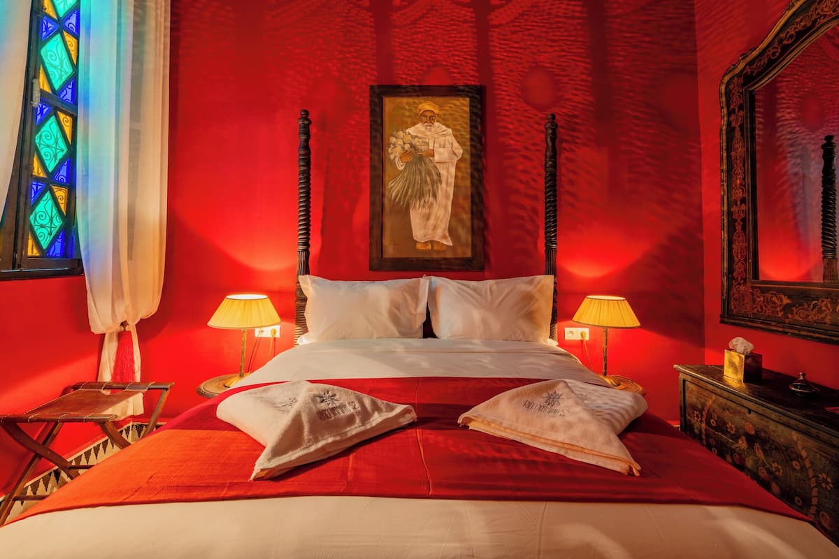 A hotel room with a red wall, a bed with white linens, two bedside lamps, a framed picture on the wall, and a small table with a lamp.