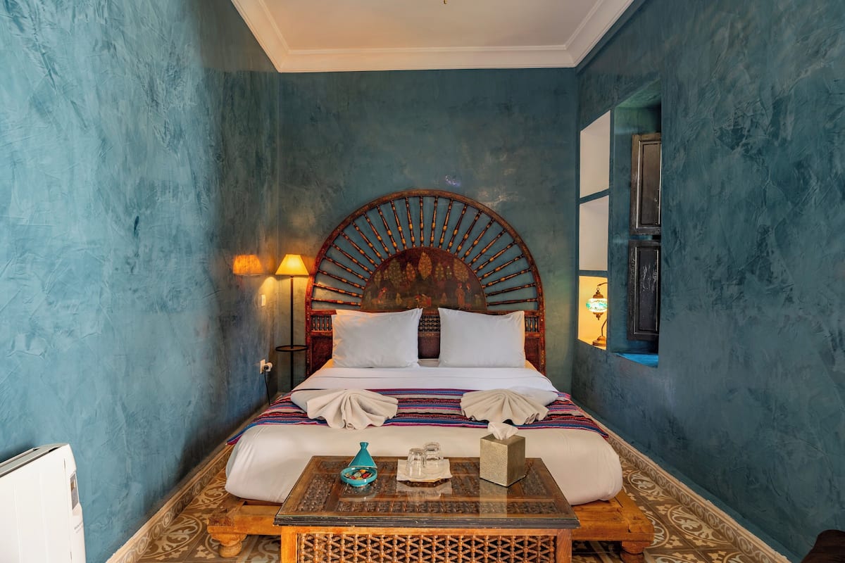 A room with a large bed, a wooden headboard, a bedside table with a vase, and a wall-mounted lamp.