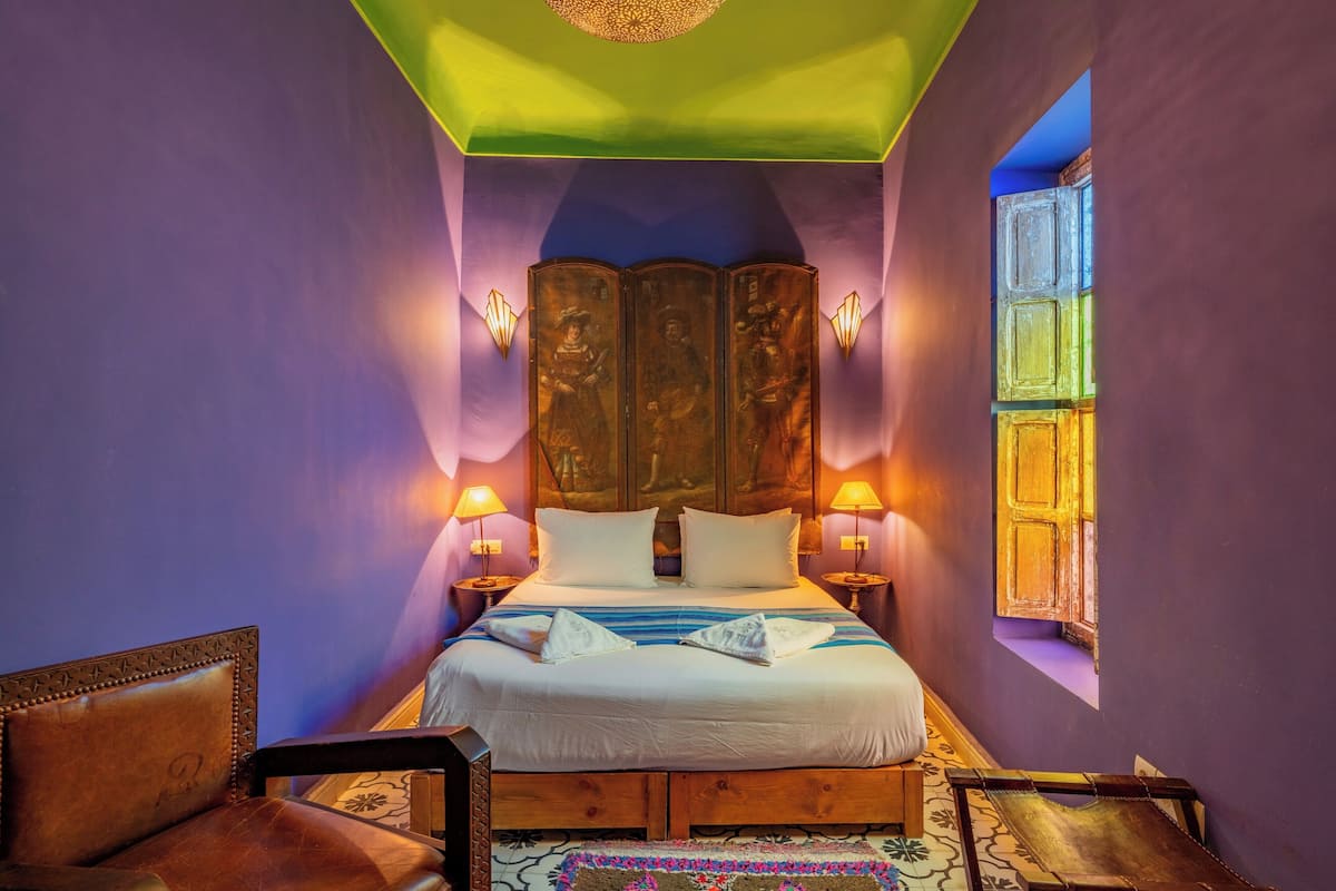 A room with a large bed, two bedside lamps, a wooden headboard, and a window with a wooden door.