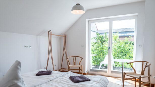 A modern bedroom with a bed, two wooden chairs, a balcony with a view of greenery, and a hanging light fixture. - Gruppenhaus Darmstadt (Darmstadt)