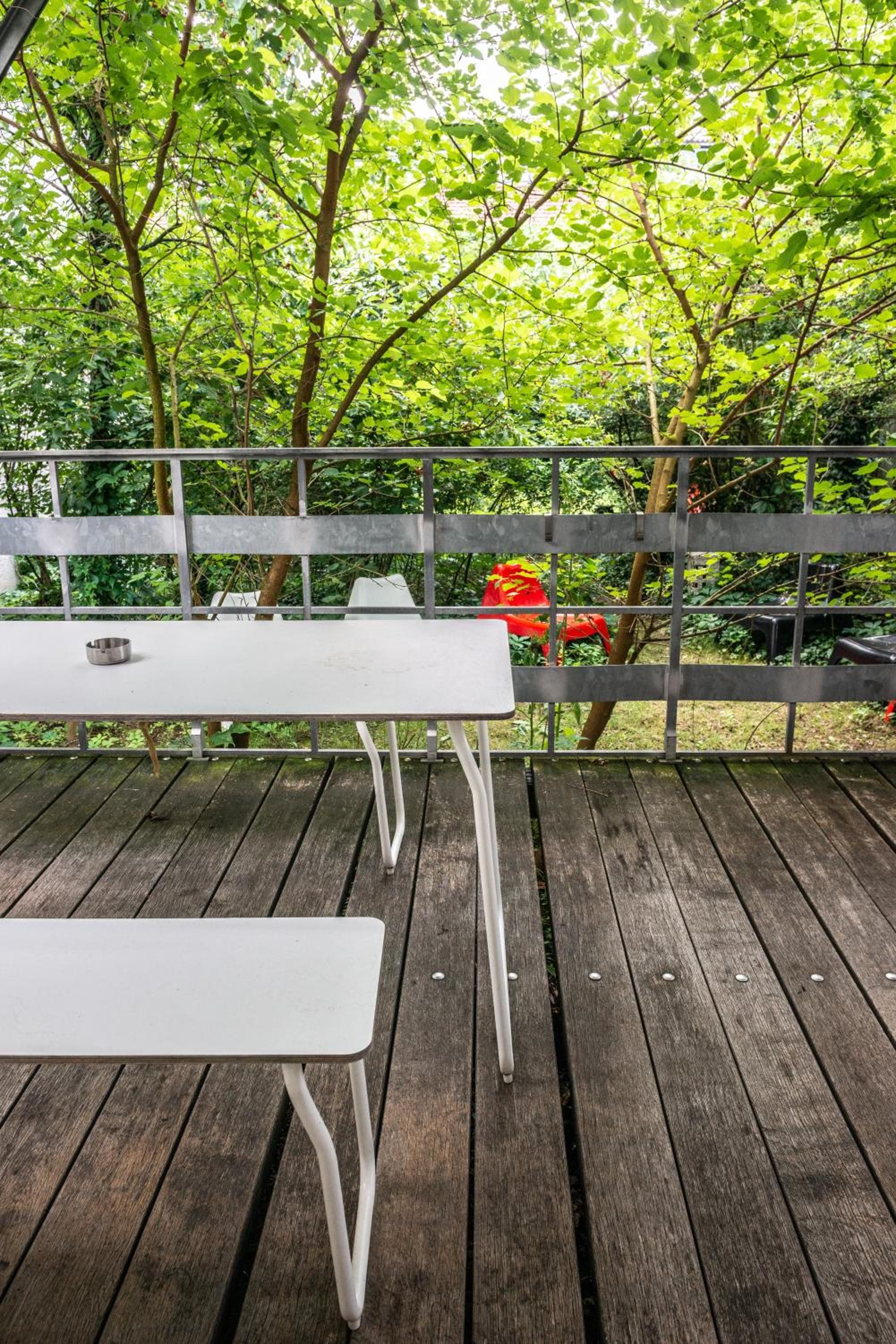 Eine Holzterrasse mit einem weißen Picknicktisch und Bänken, umgeben von üppigem Grün und einem Metallgeländer.