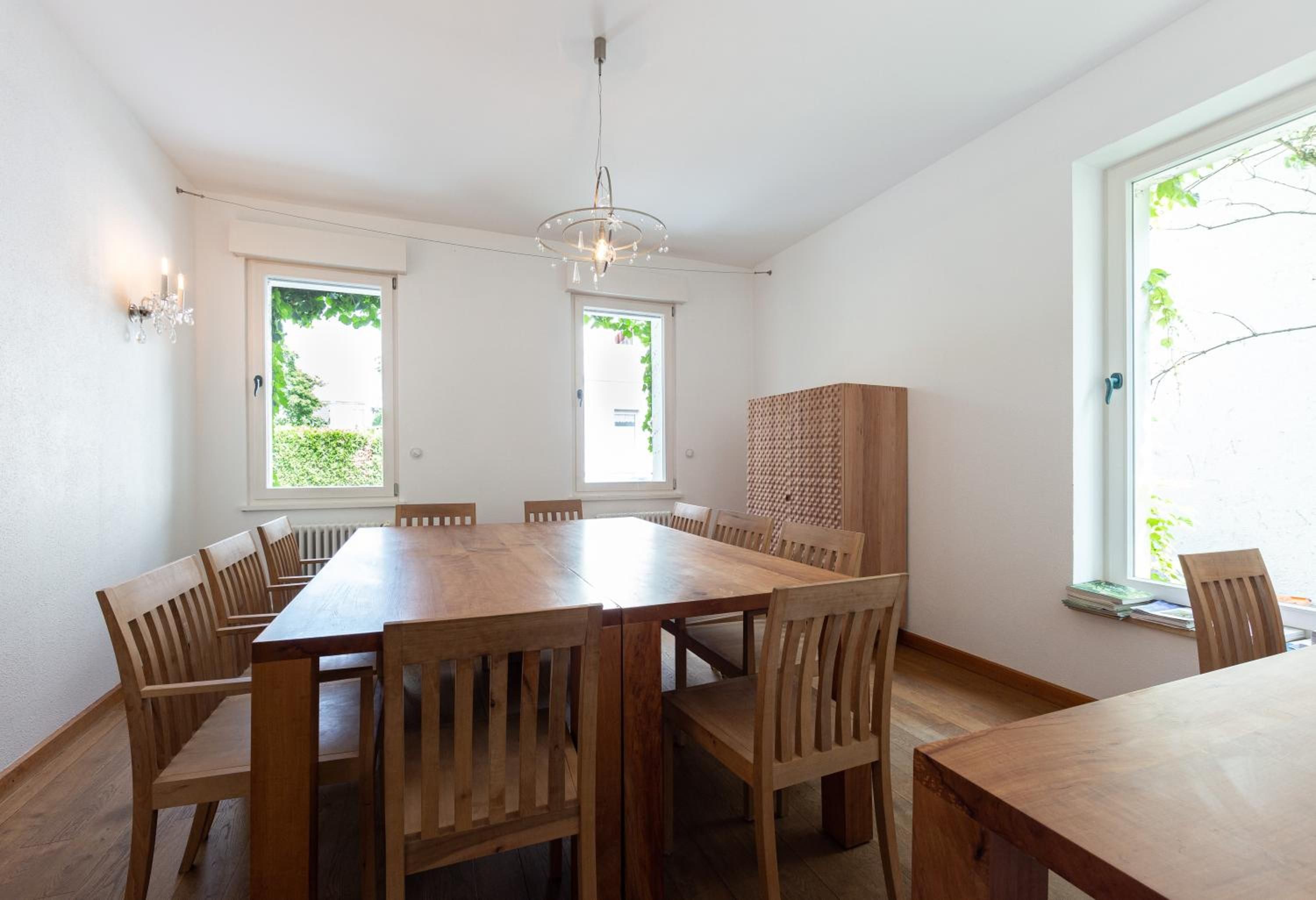 Ein Esszimmer mit Holztisch und Stühlen, einer Chandelüster und zwei Fenstern, durch die sich Grünpflanzen erkennen lassen.