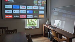 A conference room with a large screen displaying various app icons, a whiteboard, a wooden desk, and a window with books. - Gruppenhaus Darmstadt (Darmstadt)