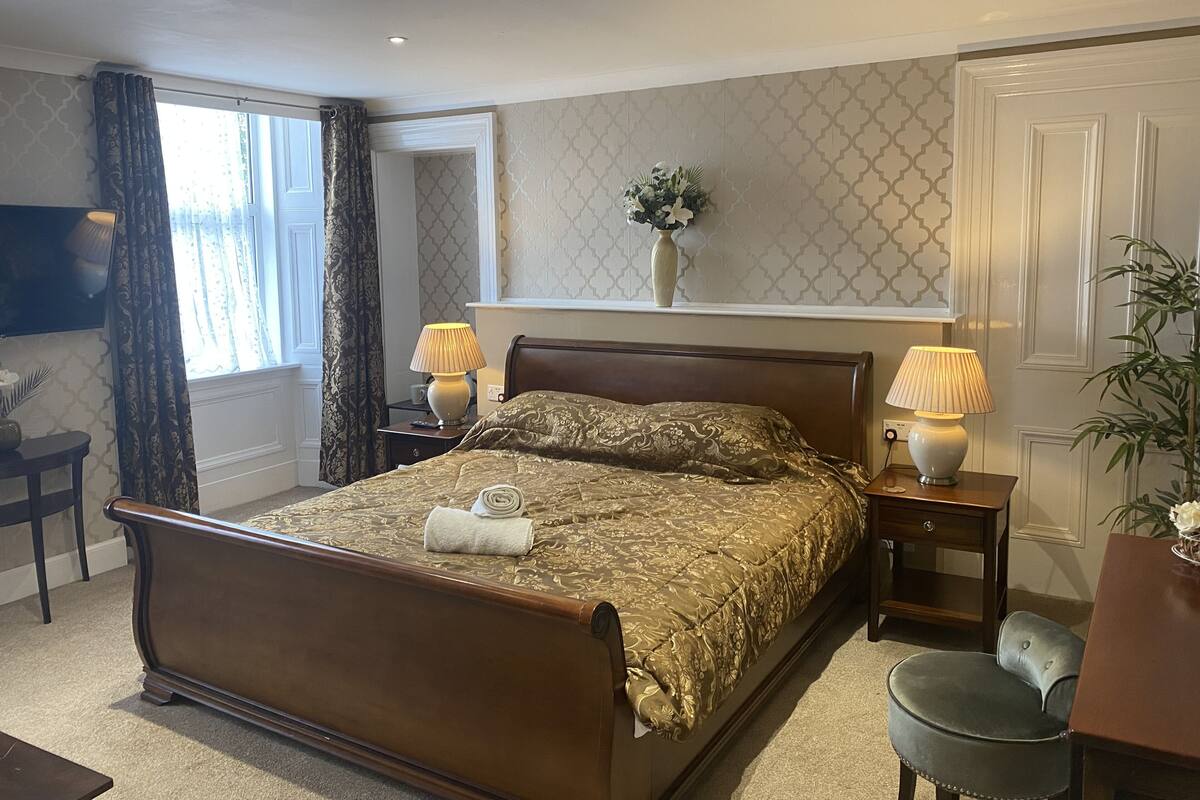 Une chambre d’hôtel bien aménagée, avec un grand lit, deux tables de chevet avec des lampes, une petite table avec une chaise, une plante en pot et un téléviseur fixé au mur.