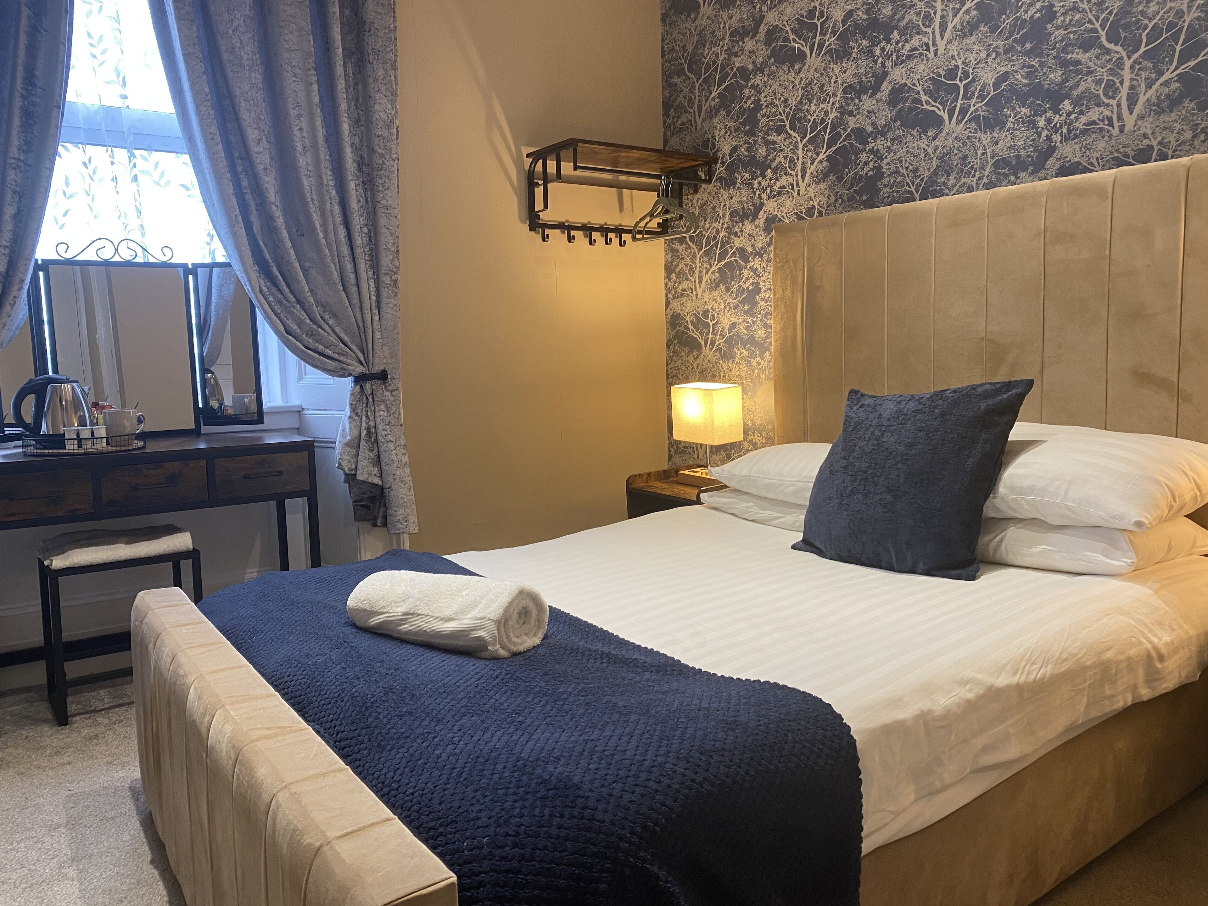 Una cama bien hecha con una manta y almohadas azules, una lámpara de noche, una mesita de noche de madera, un escritorio con una tetera y tazas, y una ventana con cortinas.