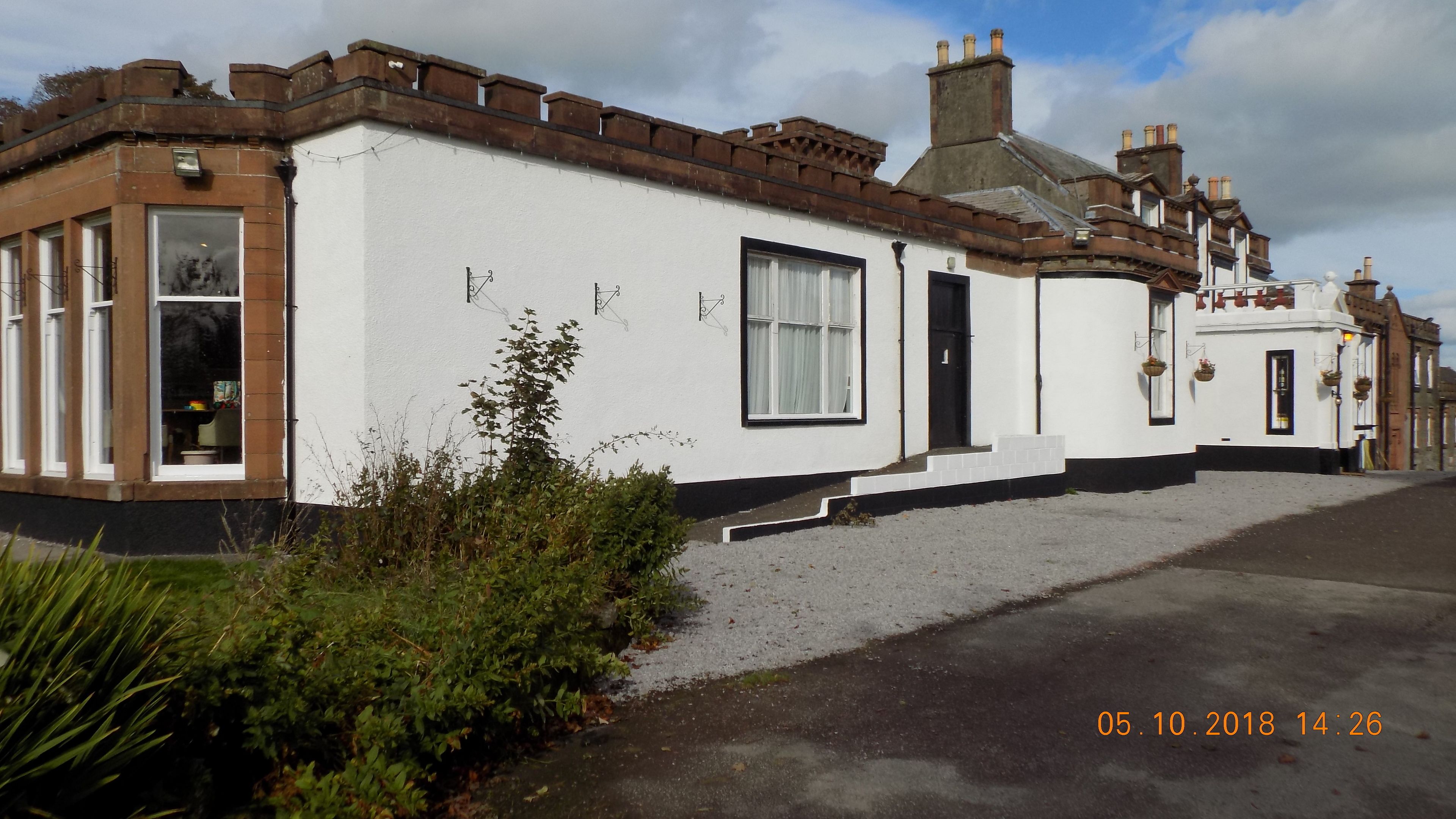 A white building with a brown roof, a black door, and a window with white curtains. There is a small garden in front.