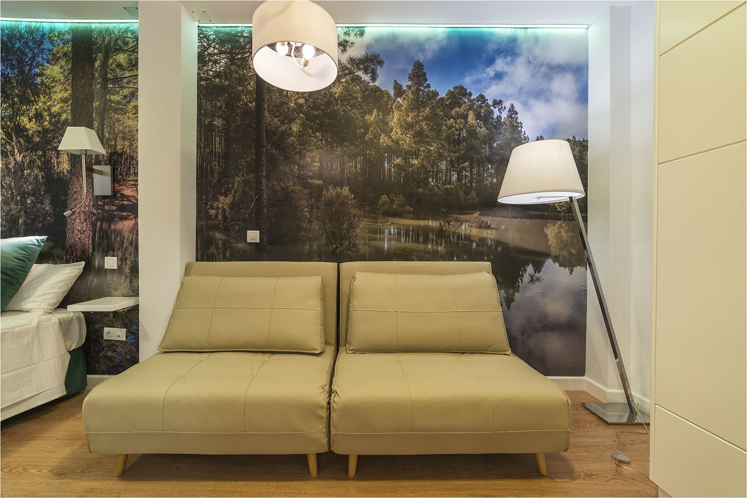 Una habitación con sofá, dos lámparas y un mural en la pared que muestra un bosque y un lago.