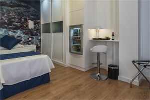 A hotel room with a bed, a minibar, a stool, and a trash bin.