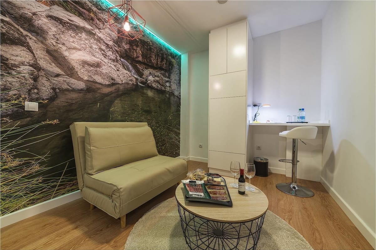 A room with a sofa, a small round table with a book and a bottle, a wall with a rock-like texture, a cabinet, and a desk with a chair.
